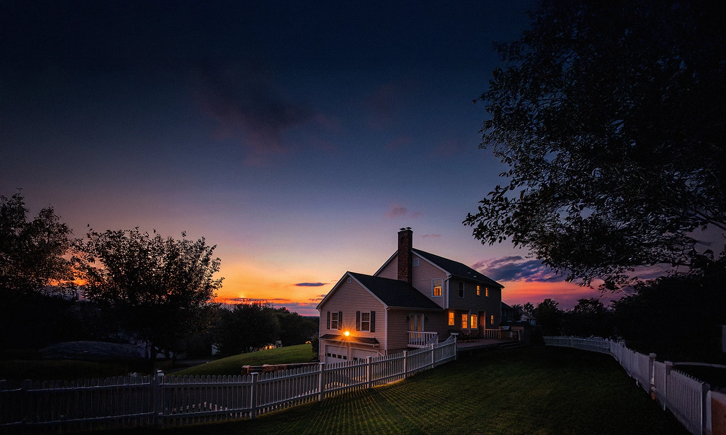 Beautiful house at twilight with warm lights glowing from windows, surrounded by white picket fence and green lawn under a dramatic sunset sky